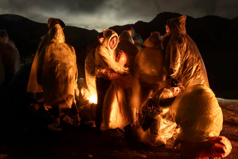 A group of people wearing translucent rain ponchos huddle together at night on a rocky terrain, illuminated by a warm light against dark, mountainous surroundings. Some people appear to be comforting each other.