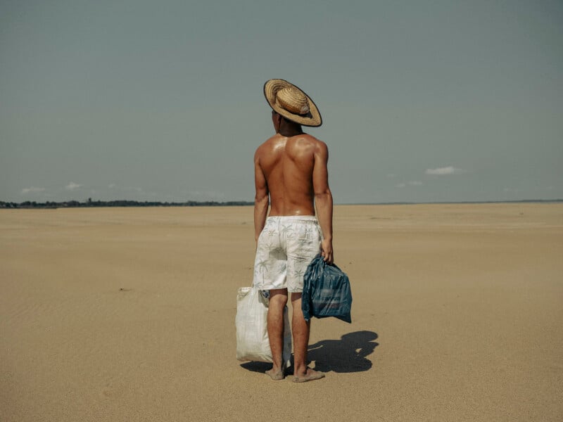 A shirtless man wearing a straw hat and white shorts stands barefoot on a vast sandy plain, holding two bags, one in each hand, and looks toward the distant horizon under a clear sky.
