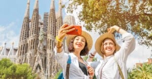 Two people in hats take a selfie with a smartphone in front of the Sagrada Família in Barcelona. They are smiling and appear to be enjoying a sunny day, with trees and the iconic church spires in the background.
