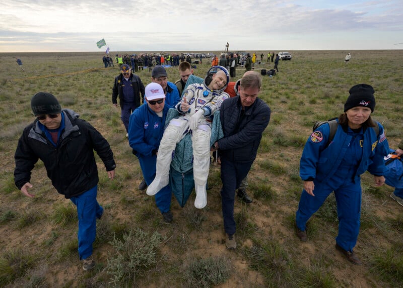 A group of people in blue jumpsuits and jackets carry an astronaut in a white spacesuit across a grassy field, with a crowd and vehicles in the background under a cloudy sky.