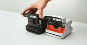 A hand adjusts two instant cameras on a white surface. The cameras are side by side, one black and the other white with an orange accent. Both have visible lenses, viewfinders, and flash units.