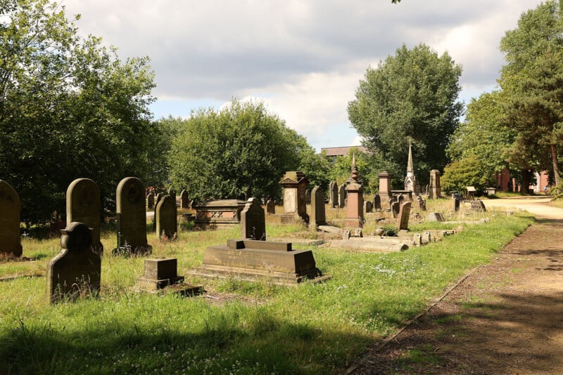 A sunlit cemetery with various old gravestones and monuments surrounded by green grass, trees, and a dirt path on the right, under a partly cloudy sky.