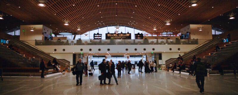 Spacious train station interior with high arched ceiling and warm wood paneling. People walk with luggage on a patterned floor. Two staircases lead up to a mezzanine with "Loyal Legion Beer Hall" signage. Natural light streams through large windows.