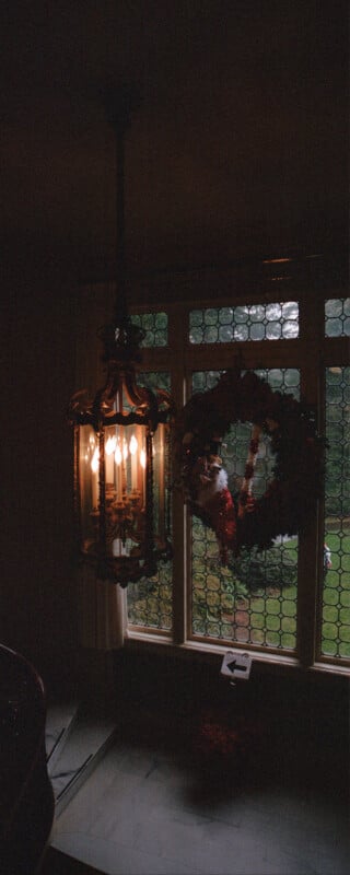 A dimly lit room features an ornate lantern emitting warm light next to a circular window. A festive wreath with red decorations and a Santa face hangs on the window. Outside, lush greenery is visible through the glass.