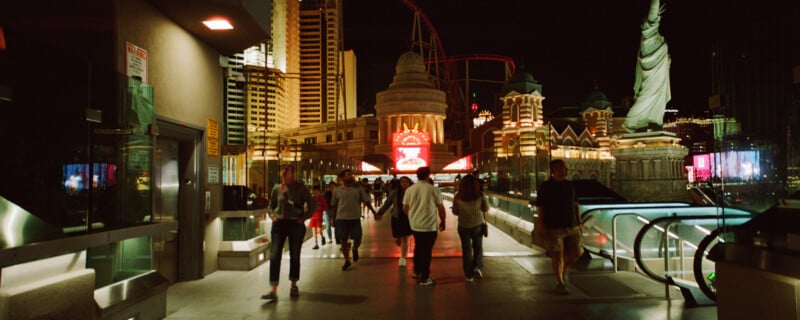 People walking on a brightly lit pedestrian walkway at night in an urban area, surrounded by illuminated buildings and a replica of the Statue of Liberty. Signs and lights create a vibrant atmosphere.