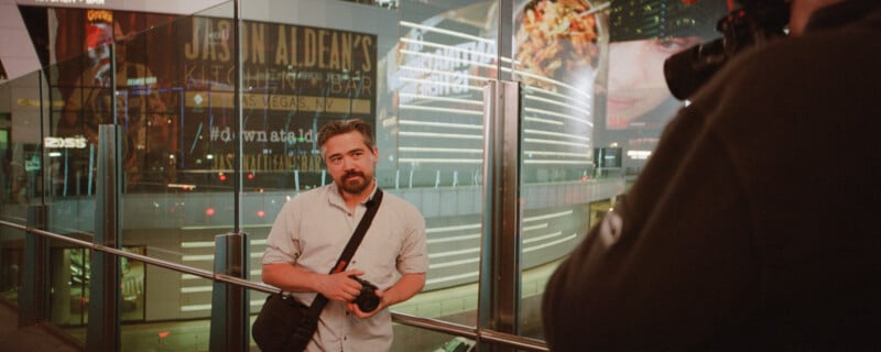 A man with a beard holds a camera and poses for a photo in an urban setting at night. Neon lights and advertisements illuminate the background. Another person is partially visible, holding a camera in the foreground.