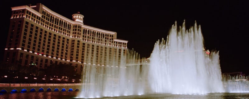 A large hotel with bright lights is seen at night behind a display of water fountains shooting high into the air over a pond below. The hotel has a curved structure and multiple floors illuminated by lights.