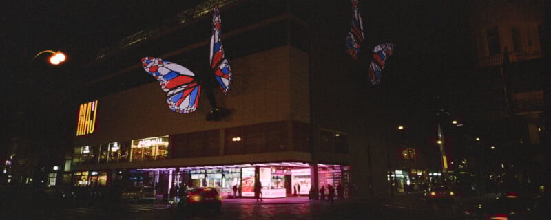 Night scene of a brightly lit storefront with illuminated butterfly decorations. The building displays "H&M" in large letters. Various signs and people are visible on the bustling street, with cars passing by.