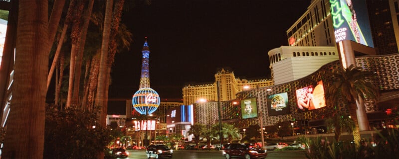 Night view of a bustling street in Las Vegas, featuring palm trees, neon signs, and bright lights. The faux Eiffel Tower stands prominently amid illuminated casinos, billboards, and cars. The atmosphere is vibrant and lively.