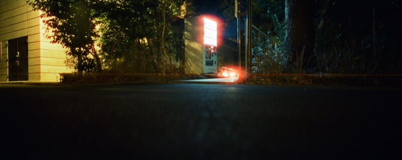 Low-angle nighttime image of a dimly lit street corner featuring a glowing sign, surrounded by trees and foliage. A car's red brake lights are visible in the distance, casting a faint reflection on the pavement.