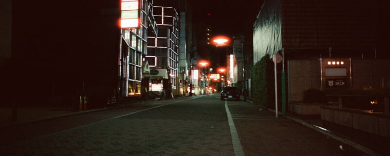 Nighttime urban street scene with illuminated neon signs and streetlights lining a mostly empty road. Buildings have modern, geometric designs. A few parked cars are visible in the distance. The atmosphere is calm and quiet.
