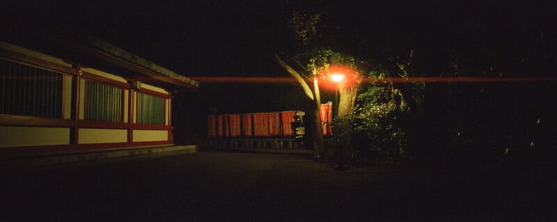 Dimly lit scene showing a path alongside a traditional Japanese building at night. A red lantern casts a warm glow on nearby trees and shrubbery. In the background, red torii gates are partially visible, creating a serene and atmospheric setting.