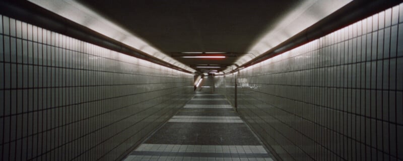A narrow, empty underground hallway with tiled walls and a tiled floor stretches into the distance. Overhead lights cast a dim glow, creating a sense of depth and perspective.