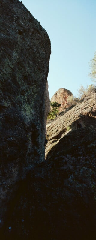 A climber scales a rocky cliff, partially shaded with sunlight hitting the upper rocks. The sky is clear and blue, with a few trees visible on the right.