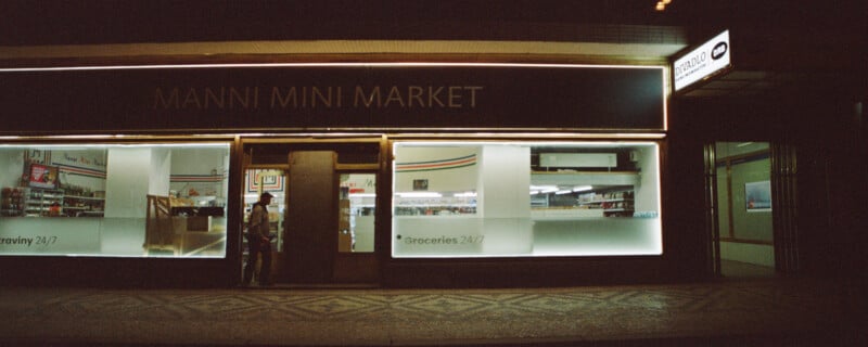 Street view of a small convenience store at night with illuminated signs. The name "Manni Mini Market" is displayed above, along with "Groceries 24/7" on the windows. The interior is visible through the glass, showing aisles and a person inside.