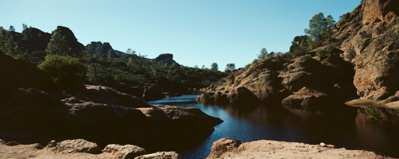 A serene landscape featuring a calm river winding through rocky terrain with sparse green foliage. The clear blue sky contrasts with the rugged, earthy rock formations and tranquil water, creating a peaceful natural scene.