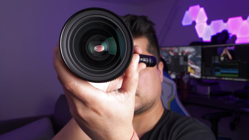 A person holds a large camera lens close to their face, partially obscuring it. In the background, a computer monitor and geometric LED lights are visible on the wall.