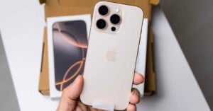 A person holds a gold smartphone with three camera lenses on the back, featuring the Apple logo. The phone is positioned over an open box, with another phone visible inside. The setting appears to be a table or desk.