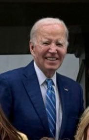 An older man with short white hair, wearing a blue suit, light blue shirt, and blue striped tie, stands smiling outdoors.