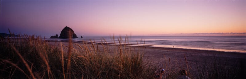 A beach scene at sunset with calm waves and a large rock formation in the distance. The sky transitions from soft pink to purple hues. In the foreground, tall grass borders the view.