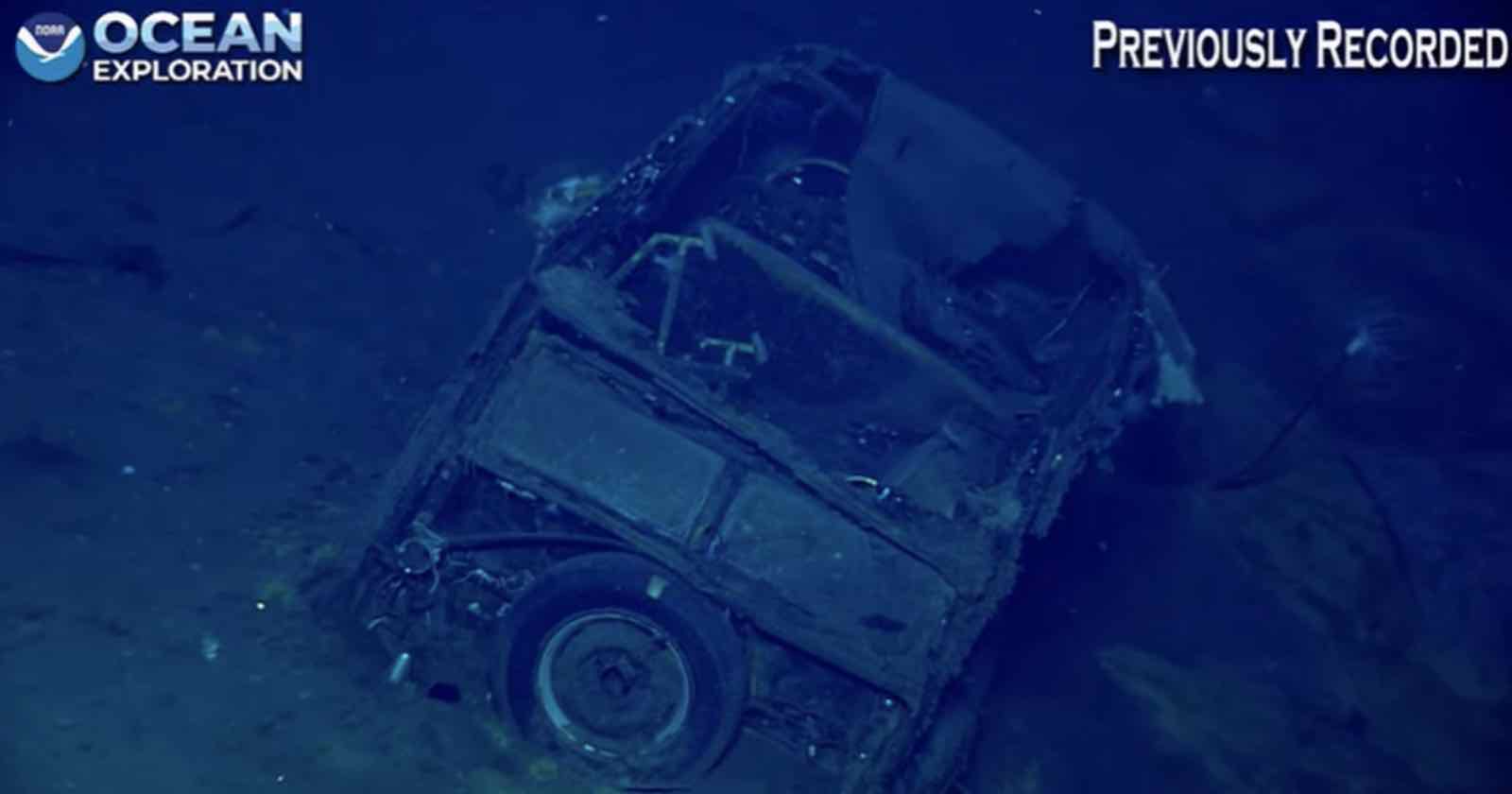 A rusted, partially buried vehicle rests on the ocean floor, captured in deep blue water with "Ocean Exploration" and "Previously Recorded" labels on the image.