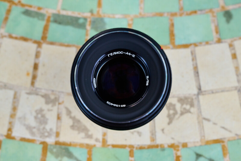 A black camera lens with Cyrillic writing is photographed from above, centered against a tiled background of alternating green and beige squares.