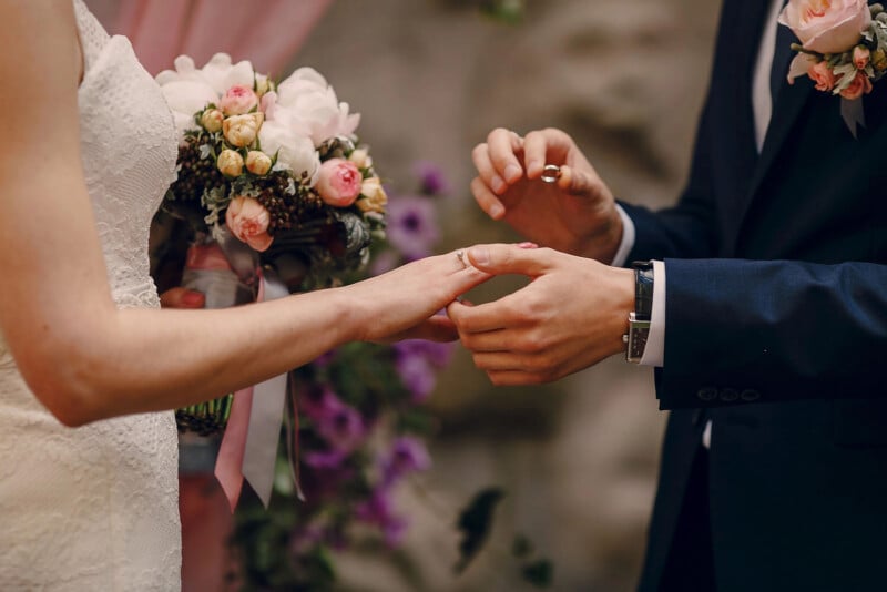 A groom in a dark suit places a ring on the bride's finger during a wedding ceremony. The bride holds a bouquet of pink and white flowers, and both are dressed formally. Flowers and decorations are in the background.