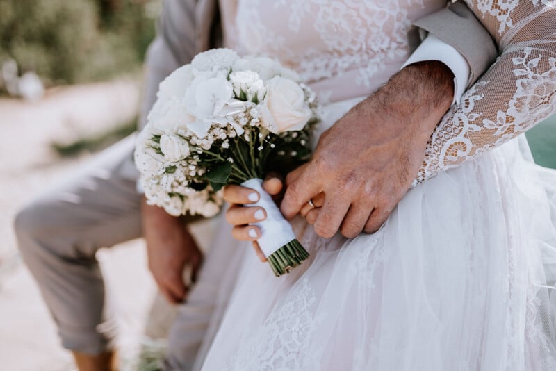 Close-up of a bride and groom. The bride holds a bouquet of white flowers, and both hands are intertwined. She wears a lace wedding dress, and he wears a light suit. Their faces are not visible.