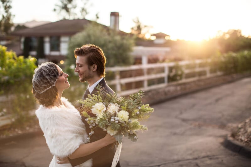 A bride and groom smile at each other outdoors at sunset. The bride wears a white fur shawl and holds a bouquet, while the groom wears a suit. Soft, golden sunlight creates a romantic atmosphere.