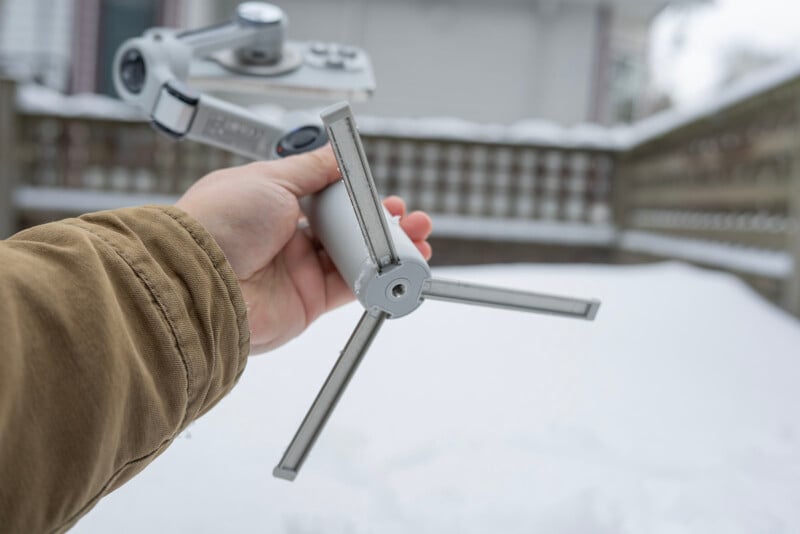 A person wearing a brown jacket holds a foldable camera tripod with a smartphone mount in a snow-covered outdoor setting. The tripod is partially extended, and a wooden fence and house are visible in the background.