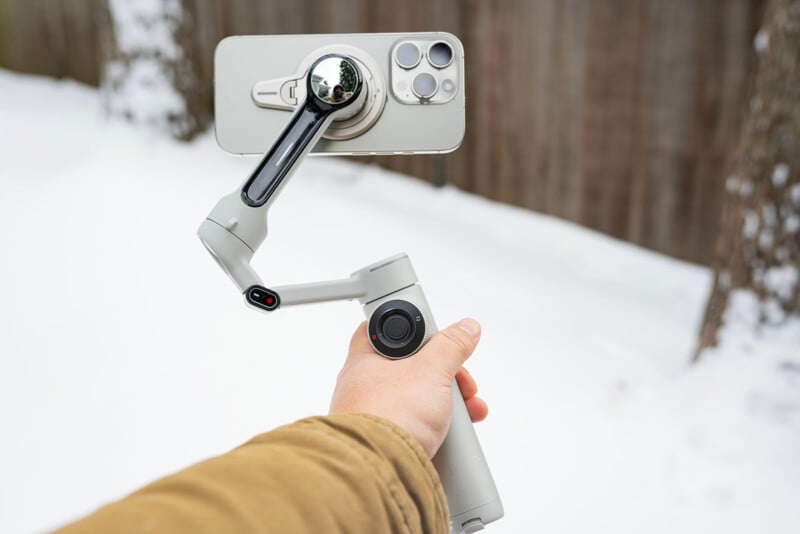 A person holding a smartphone mounted on a gimbal stabilizer in a snowy outdoor setting. The phone screen is facing outward, and the person is wearing a brown jacket. The gimbal is white with black accents.