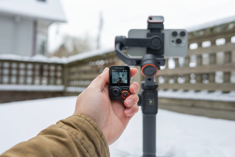 A person holds a handheld camera with a small screen displaying a snowy outdoor scene. In the background, a smartphone is mounted on a stabilizer. The setting is a snow-covered patio with a wooden fence and a house visible.