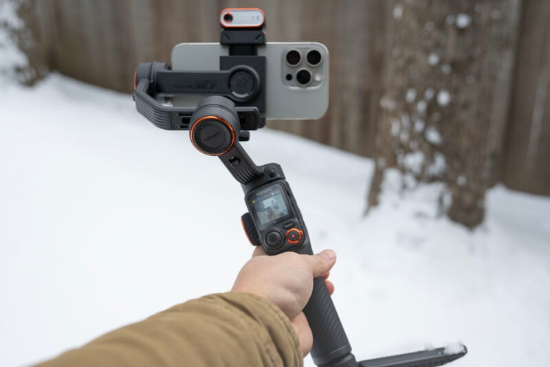 A person's hand holding a smartphone mounted on a handheld gimbal, with a snowy outdoor background. The gimbal has a small screen and control buttons. The phone has an attachment on top.