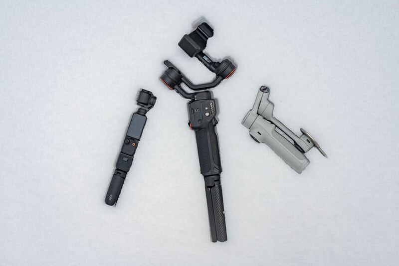 Three camera stabilization gimbals are arranged on a light surface. Two are handheld, with black and dark gray elements, and one is a compact, foldable model with a light gray body.