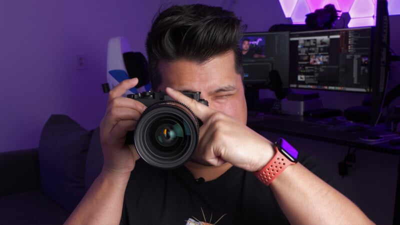 A person with dark hair is holding a camera up to their face, taking a photo. They are indoors, wearing a red smartwatch, with computer monitors and equipment visible in the background.