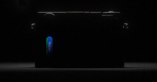 A dark, low-light image of a camera, mostly in shadow, with a bright blue and green label reading "PROVIA" illuminated on its side. The camera's outline and dials are just barely visible.