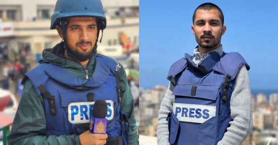 Two journalists wearing blue PRESS vests. The one on the left, in a helmet, holds a microphone with a purple and yellow logo, standing in front of a blurred cityscape background. The one on the right, without a helmet, stands against a backdrop of buildings and sky.
