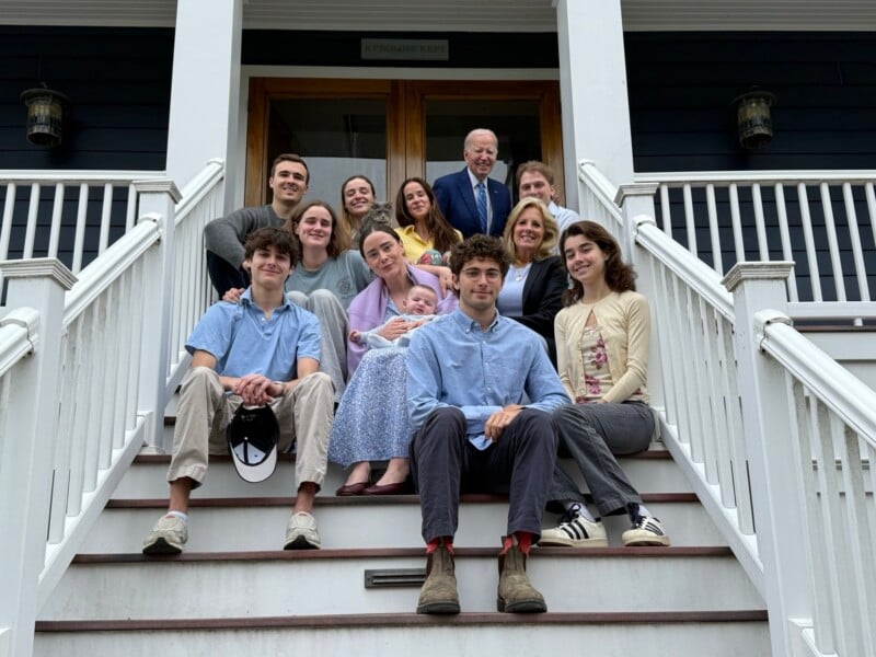 A group of thirteen people, including men, women, and a baby, pose together on the steps of a house with white railings and a wooden door, smiling for the camera.