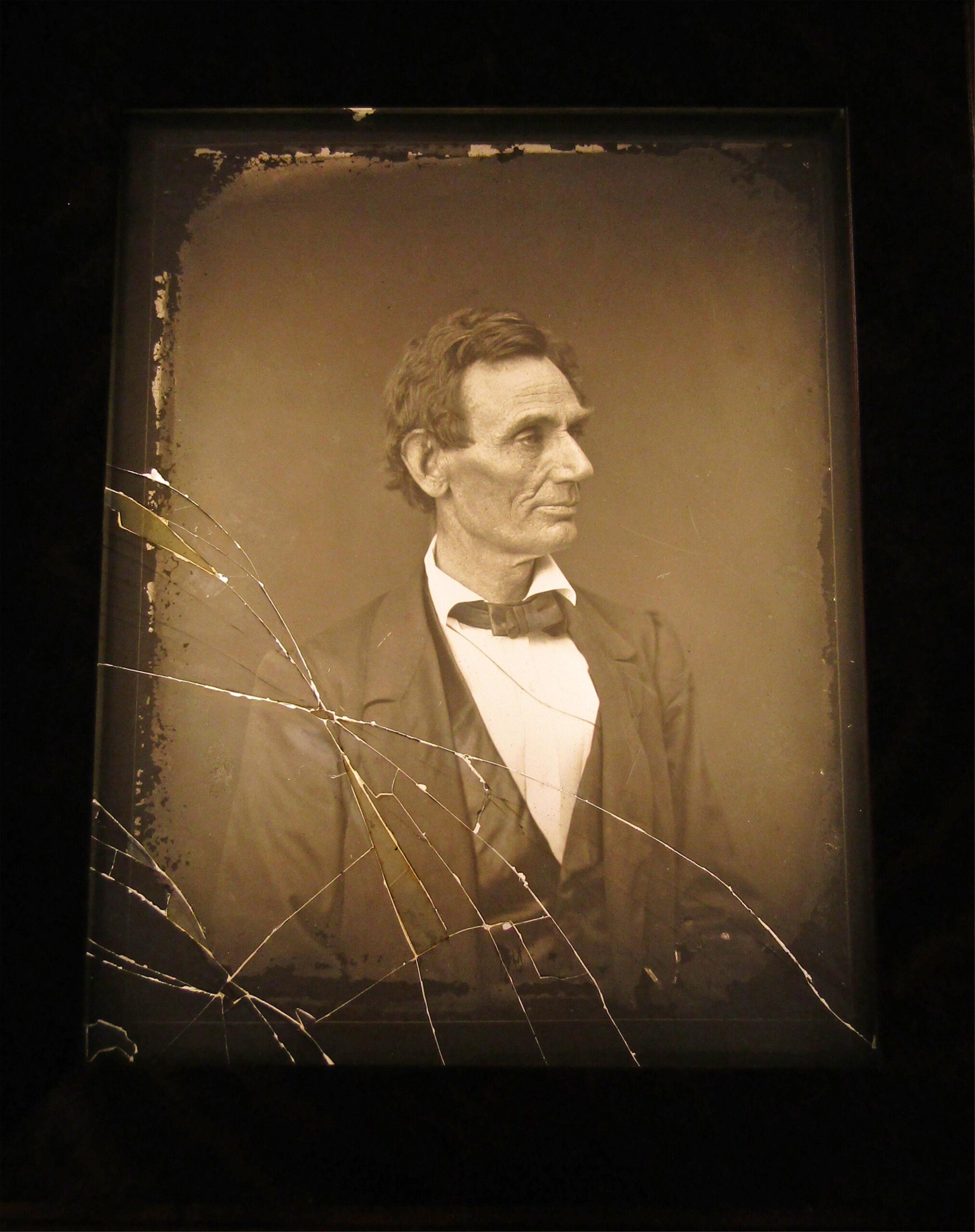 A sepia-toned, cracked and damaged photograph of a man in a suit with a bowtie, gazing to the right. The image has a vintage, worn appearance, suggesting it is from the 19th century.