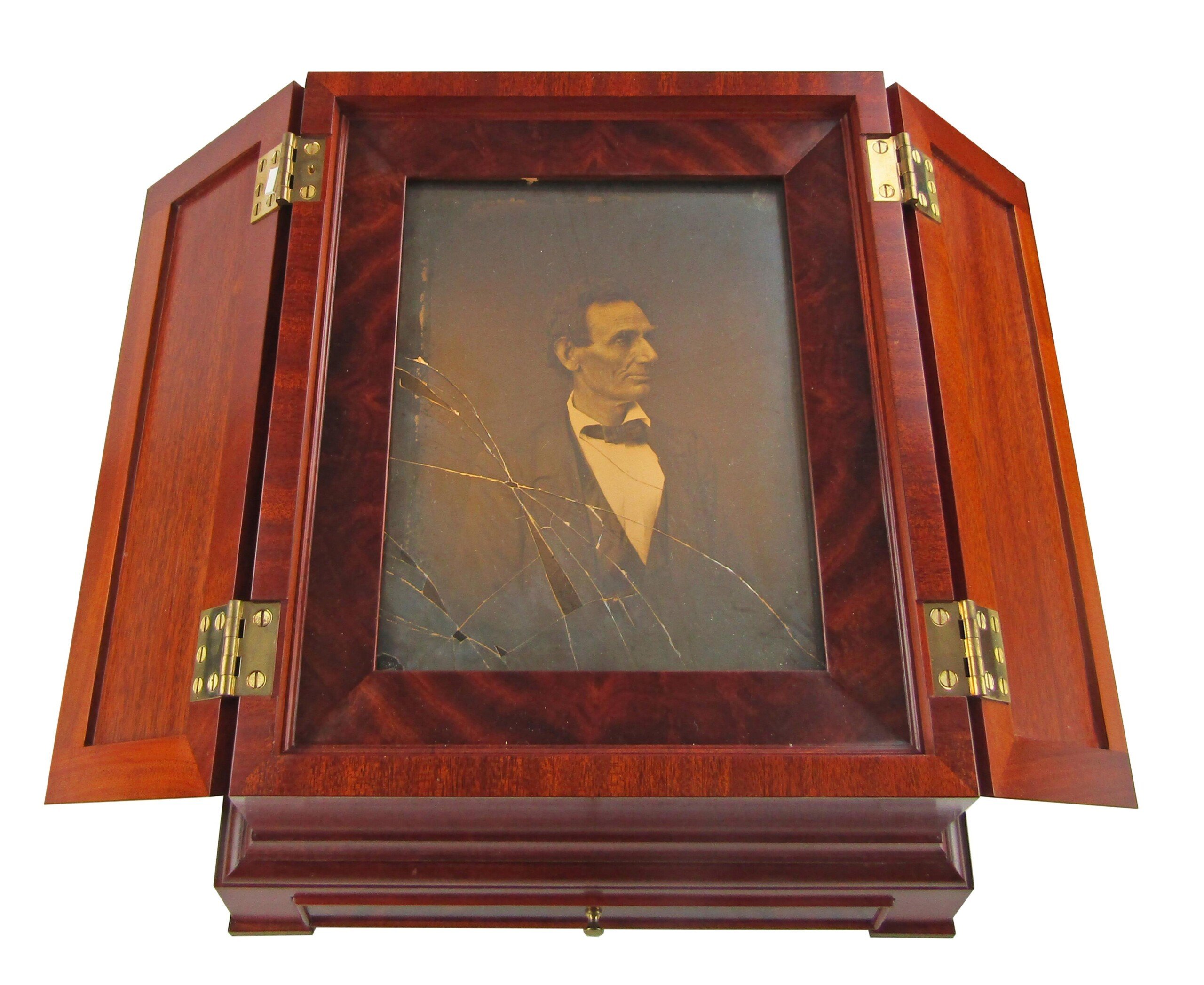 A framed, vintage black-and-white portrait of Abraham Lincoln is displayed in a wooden case with open hinged doors. The photograph glass is cracked, and the wooden case has a polished, reddish finish with brass hinges.