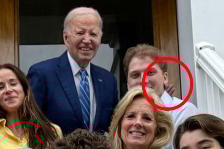 A group of people poses on steps for a photo. An older man in a blue suit stands near the center, smiling. One man in the back is partially obscured and circled in red. Others are gathered around, also smiling.