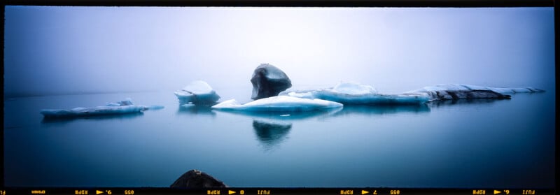 A tranquil scene of icebergs floating on calm water, under a foggy sky. The icebergs are various shapes and sizes, with reflections visible in the water, creating a serene and peaceful atmosphere.