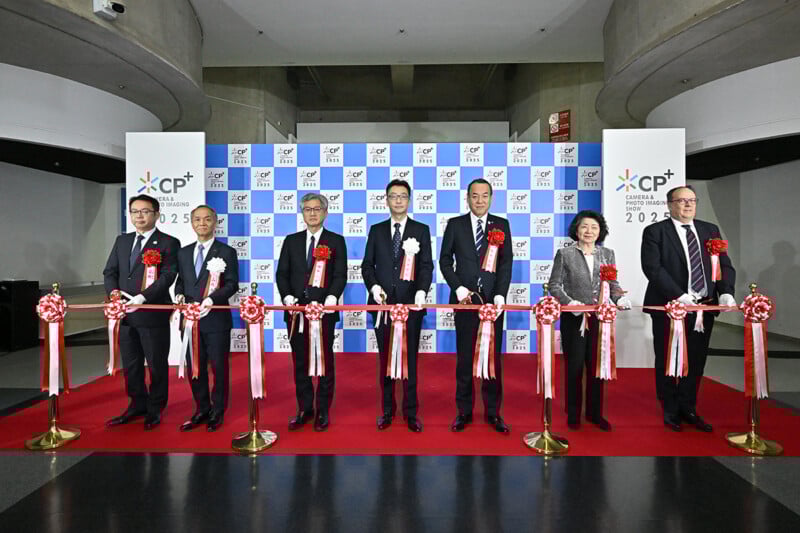 Seven people in formal attire stand on a red carpet, holding a ribbon at a ceremony. Behind them is a blue and white backdrop with “CP+ 2025” and logos. Four men hold large scissors, preparing to cut the ribbon.