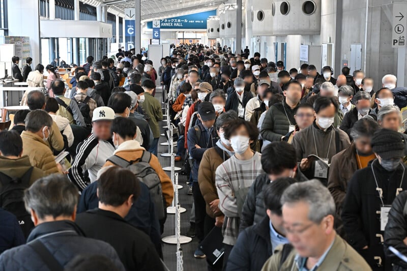A large crowd of people, many wearing masks, stand in long lines inside a spacious indoor venue with high ceilings and signs overhead. The atmosphere appears busy and organized.