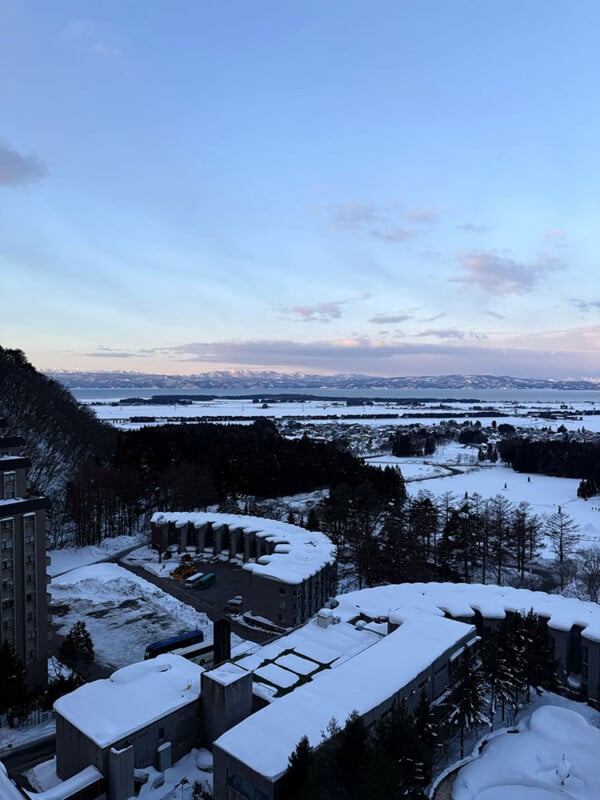 View from a high vantage point of snow-covered buildings, trees, and fields stretching into the distance, with mountains and a partly cloudy sky at dawn or dusk.