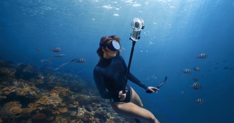 A woman in a black wetsuit is underwater, holding a camera on a selfie stick while surrounded by tropical fish and coral reefs. She is wearing a diving mask and snorkel.