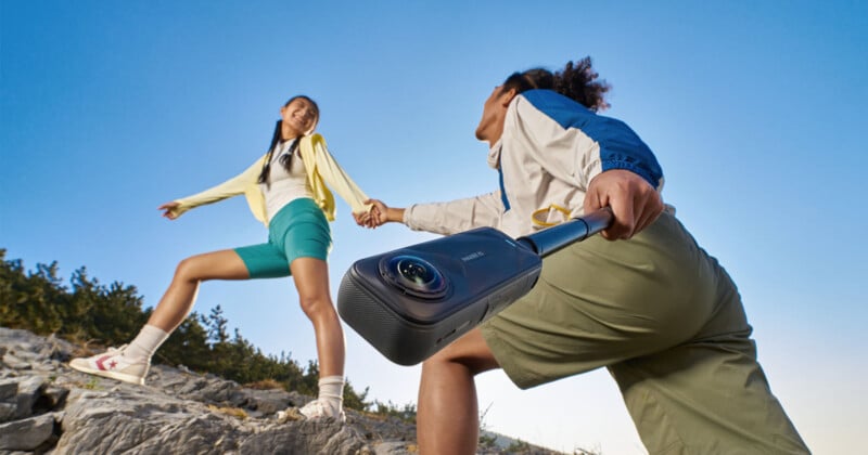 Two people hiking on rocky terrain, holding hands. One person is in the foreground holding a portable camera or device on a stick, with blue sky and trees in the background.