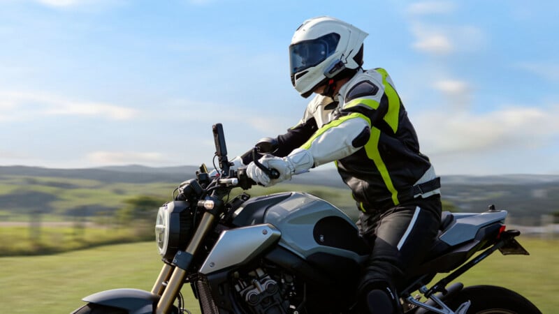 A person wearing a white helmet and black motorcycle gear with neon accents is riding a modern motorcycle outdoors, with green hills and a blue sky in the background.