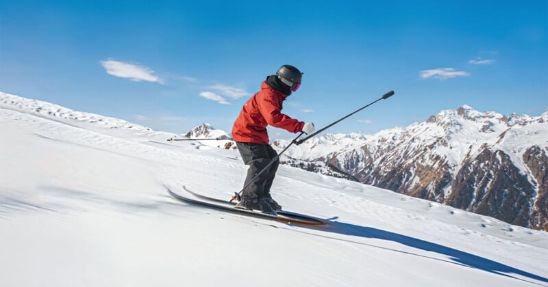 A skier wearing a red jacket and helmet glides downhill on a snowy mountain slope, holding ski poles. Snow-covered peaks and a clear blue sky are visible in the background.