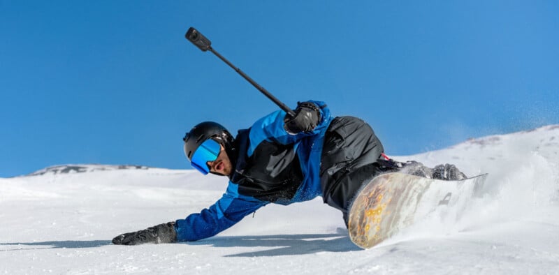 A snowboarder in blue and black gear, wearing a helmet and goggles, carves a sharp turn on a snowy slope while holding a camera on a pole under a clear blue sky.